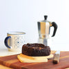 Eggless Almond Fudge Brownie topped with roast almonds on a wooden board with a coffee machine in the background.