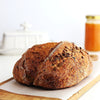 Loaf of  sourdough bread with seeds on a wooden cutting board with a jar of jam in the background.