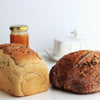 Two loaves of bread with a jar of jam and a teacup in the background on a light surface.